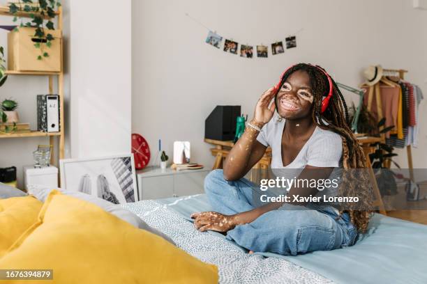 happy african woman with wireless headphones enjoying online music or podcast sitting on bed at home. entertainment and leisure concept. - vitiligo stock-fotos und bilder