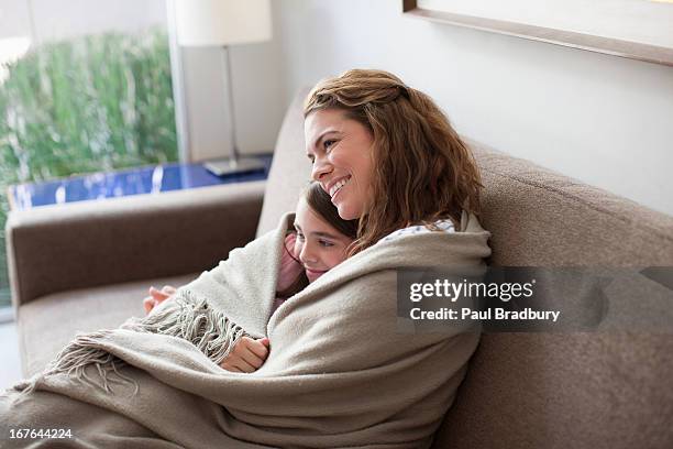 mother and daughter wrapped in blanket on couch - gezellig stockfoto's en -beelden