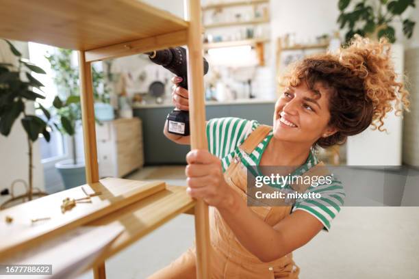 jeune belle femme habillée avec désinvolture assemblant une étagère avec perceuse électrique à la maison - bricolage photos et images de collection