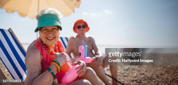 a senior couple sitting on beach loungers under an umbrella. - beach party stock pictures, royalty-free photos & images