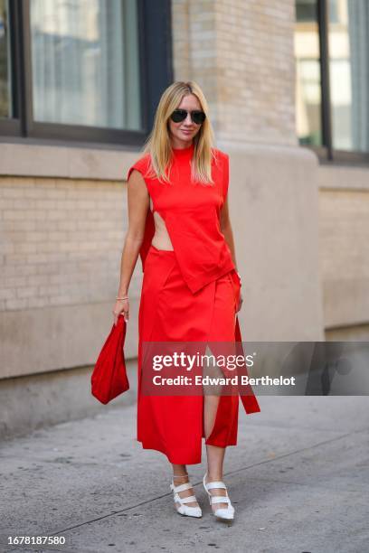 Lisa Aiken wears aviator sunglasses, a red dress made of two parts, a sleeveless top with keyhole, a red slit maxi skirt, white pointed shoes,...