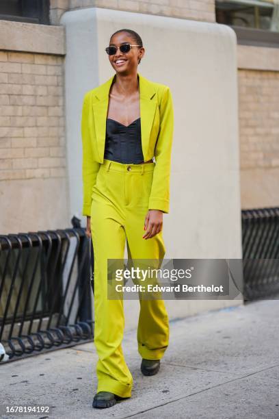 Model wears sunglasses, a black low-neck top, a neon yellow blazer cropped jacket, matching flared suit pants, black leather shoes, a bag, outside...