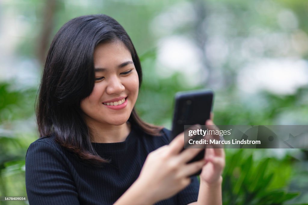 Lächelnde junge Frau, die ihr Mobiltelefon im Freien im Park benutzt