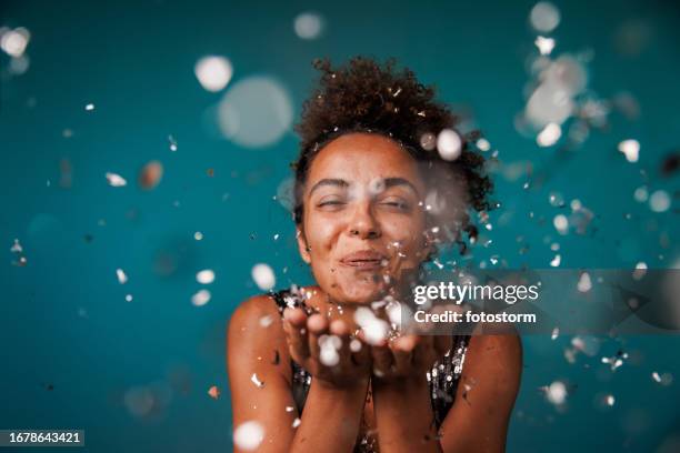portrait of joyful young woman blowing silver colored confetti at camera - celebração imagens e fotografias de stock