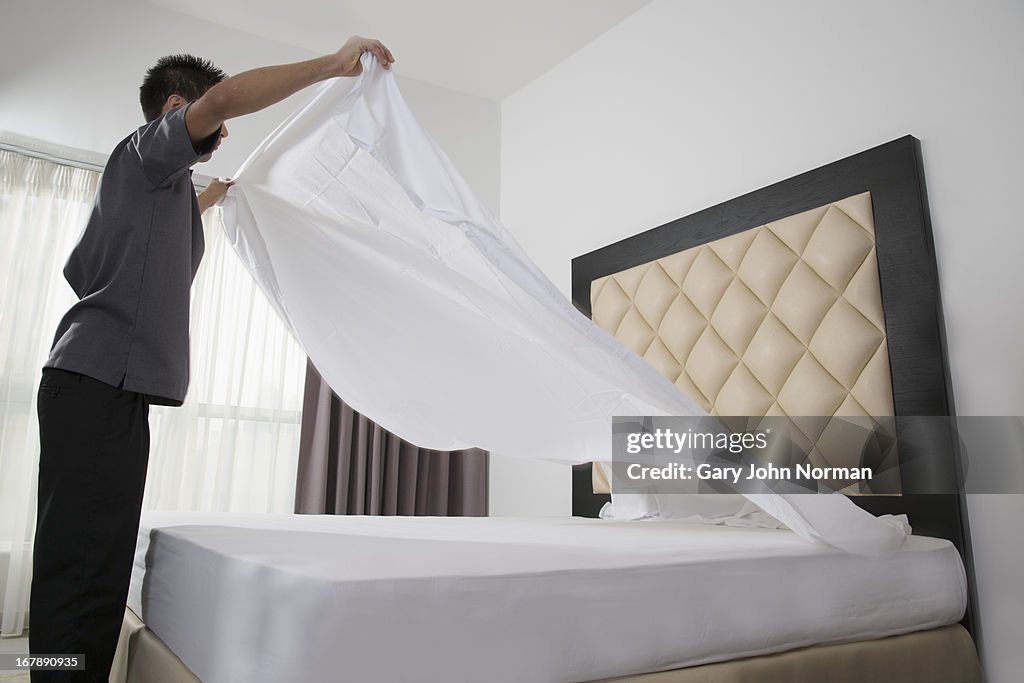 Male housekeeper in hotel throwing sheet on bed