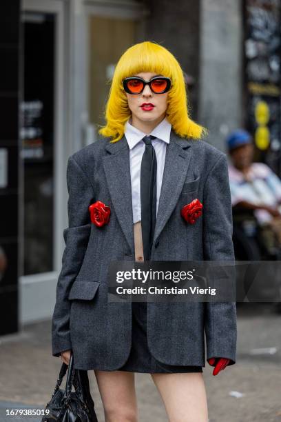 Guest with yellow hair wears grey oversized blazer, white cropped button shirt, skirt, black boots, Balenciaga bag outside Puppets and Puppets on...