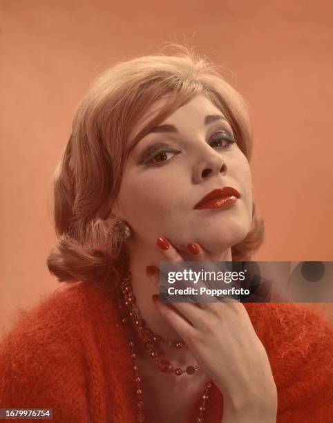 Posed studio portrait of a female fashion model wearing an orange mohair jumper and a beaded necklace, she wears blue eyeshadow, red lipstick and red...