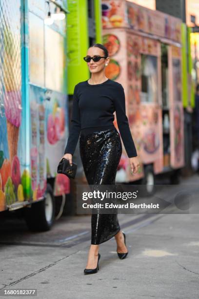 Guest wears sunglasses, earrings, a black top, a black shiny sequined skirt, pointed shoes , a clutch, outside Carolina Herrera, during New York...