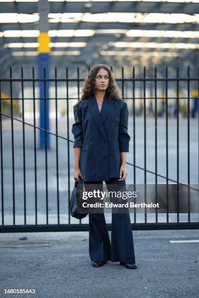 Guest wears a dark blue oversized blazer jacket, a black leather bag, dark blue striped flared suit pants, black leather shoes, outside COS, during...
