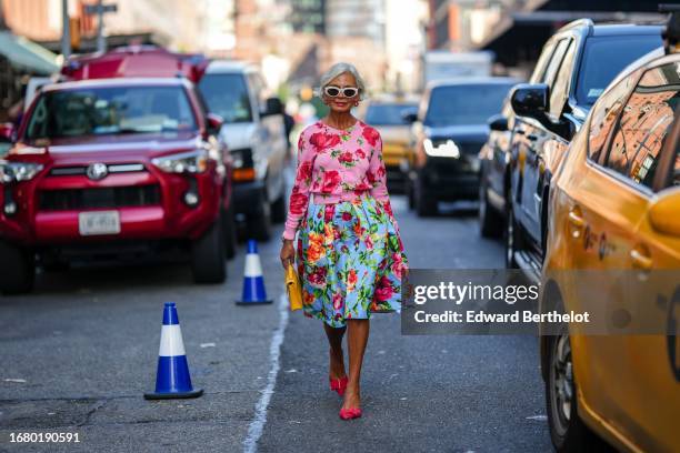 Grece Ghanem wears red earrings, sunglasses, a pink and red floral print cardigan, a blue and red floral print wide gathered on-knee skirt, red...