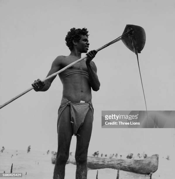 Smiling Aborigine boy stands with a stingray on his spear, his dugout canoe in the background, Arnhem Land, Northern Territory, circa 1955.