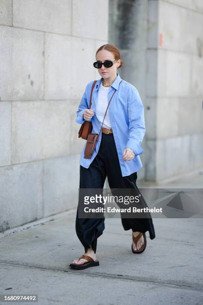 Guest wears sunglasses, a white t-shirt, a blue long oversized shirt, a brown leather bag, a belt, black palazzo flared pants, brown leather sandals,...