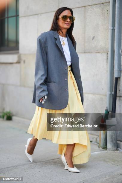Guest wears sunglasses, a golden necklace, a white t-shirt, a gray oversized blazer jacket, a pale pastel yellow pleated midi skirt, white pointed...