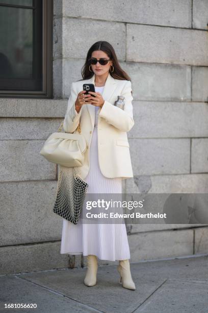 Guest wears sunglasses, earrings, a cream-colored oversized blazer jacket, a leather gathered bag, a white pleated midi dress, pointed leather boots,...