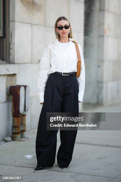 Guest wears sunglasses, a white t-shirt, a white oversized shirt, a belt, black flared slack suit pants, outside Gabriela Hearst, during New York...
