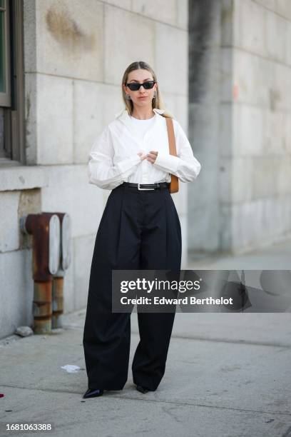 Guest wears sunglasses, a white t-shirt, a white oversized shirt, a belt, black flared slack suit pants, outside Gabriela Hearst, during New York...