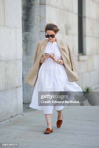 Guest wears sunglasses, a beige striped oversized blazer jacket, a white long shirt worn as a dress with embroidery, brown flat shoes, outside...