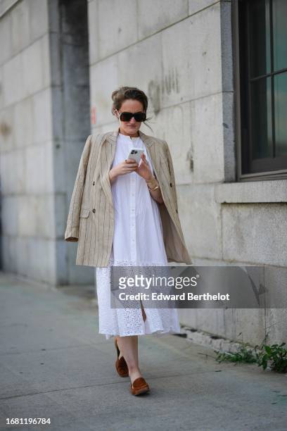 Guest wears sunglasses, a beige striped oversized blazer jacket, a white long shirt worn as a dress with embroidery, brown flat shoes, outside...