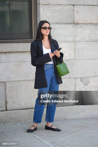 Guest wears sunglasses, a white low-neck top, a black oversized blazer jacket, blue denim jeans, a green woven leather bag, black flat ballerina...