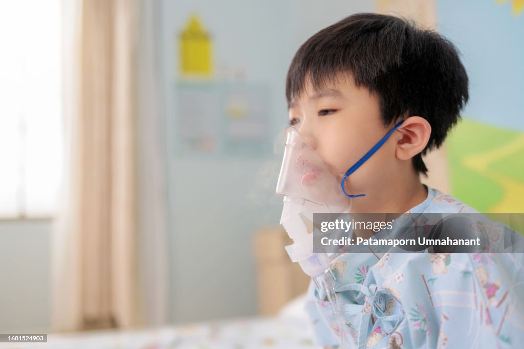 Influenza is spreading among children. Patient holding nebulizer inhaler mask, a vital tool in managing his asthma sitting on the bed in hospital ward. Deep breath through the mask make the respiratory system better. Children get the virus from school.