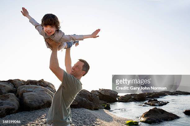 dad lifts young son above his head on beach - figlio maschio foto e immagini stock
