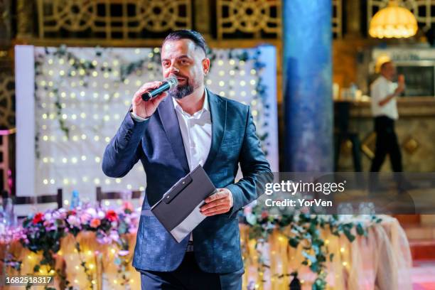 eleganter zeremonienmeister im schwarzen anzug mit mikrofon auf einer hochzeitsfeier - bridal show stock-fotos und bilder