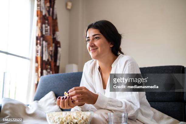 a young woman watches a movie or program on television while eating popcorn on the couch in her living room. - binge watching stock pictures, royalty-free photos & images