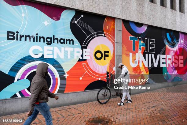 People in the city centre pass by a large scale poster advertising the city fo Birmingham as being the centre of the Universe on 21st September 2023...