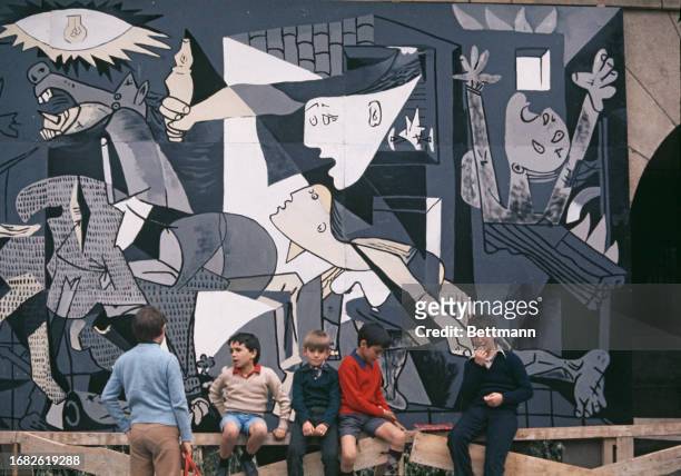 Children sitting on a fence in Fueros Square, Guernica, with a reproduction of Picasso's 'Guernica' in the background, Spain, July 28th 1977.