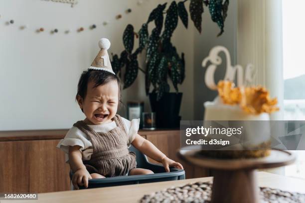 little asian baby girl wearing party hat having a tantrum while celebrating her first birthday in the living room decorated with bunting and a birthday cake at home. birthday celebration and party theme - 1st birthday stock-fotos und bilder