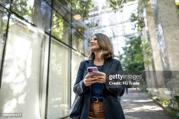 mujer de negocios madura que camina usando el teléfono móvil y mirando al aire libre - mirar alrededor fotografías e imágenes de stock