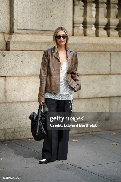 Guest wears sunglasses, a brown leather jacket, a white mesh top, black flared suit pants, a black leather bag, outside Altuzarra, during New York...