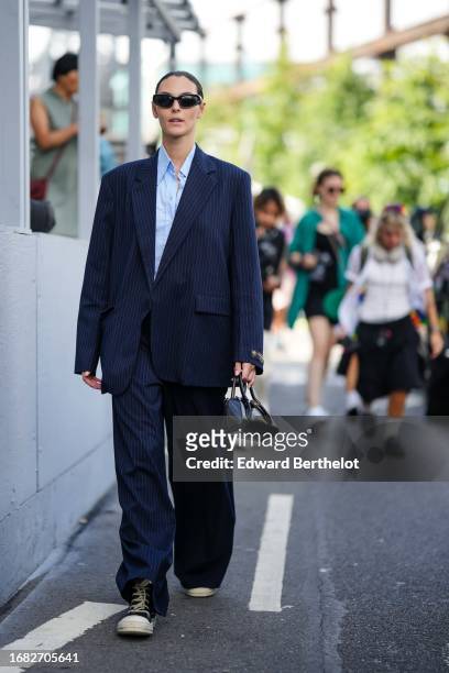 Vittoria Ceretti wears sunglasses, a blue shirt, a navy blue oversized blazer jacket, wide leg flared suit pants, sneakers, a black leather bag,...