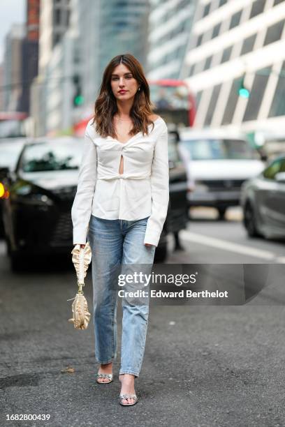 Rainey Qualley wears a white gathered low-neck top with keyhole and long sleeves, blue jeans, a golden pouch bag, silver sandals, outside Altuzarra,...