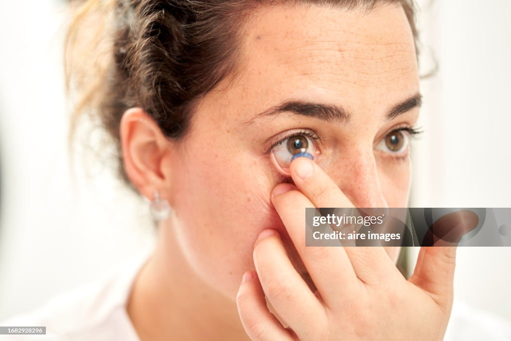 Close-up of a brown-eyed woman putting on corrective lenses in front of a mirror, white background.
