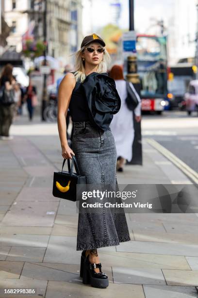 Betty Bachz wears cap, bag with banana, grey denim skirt, ruffled black blouse outside Huishan Zhang during London Fashion Week September 2023 on...
