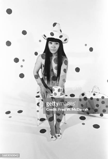 Portrait of Japanese artist Yayoi Kusama, dressed in a straw hat and shoes but otherwise naked and painted with polka dots, as she sits and holds a...