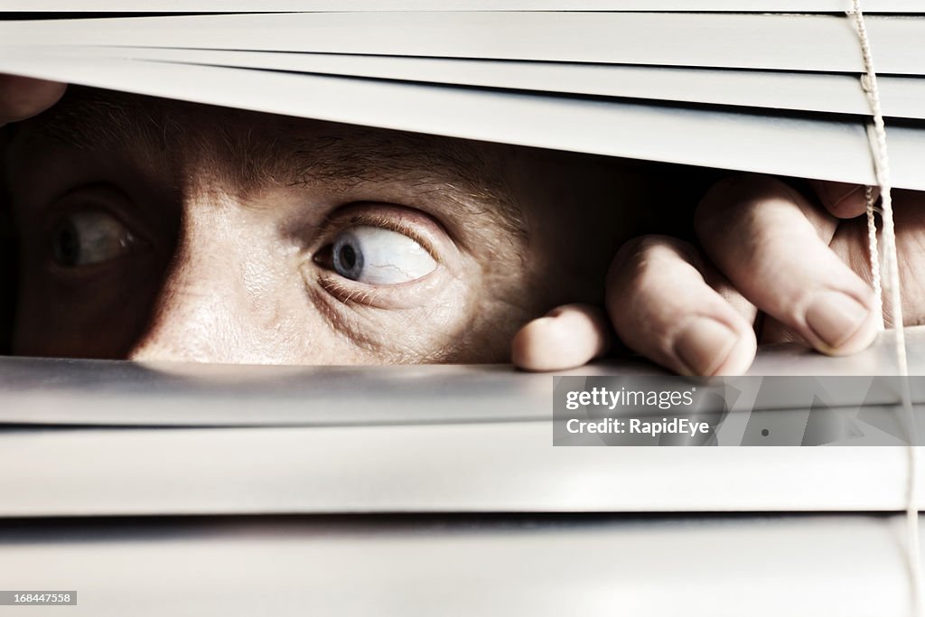 Terrified man looks to the side through venetian blinds