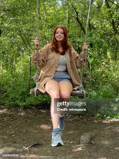 close-up image of redheaded woman wearing sunglasses on top of head, looking at camera, sitting on woodland swing in dappled sunshine, wood plank suspended from tree branch by ropes, laughing and smiling, focus on foreground - sole of shoe stock pictures, royalty-free photos & images