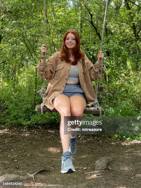 close-up image of redheaded woman wearing sunglasses on top of head, looking at camera, sitting on woodland swing in dappled sunshine, wood plank suspended from tree branch by ropes, laughing and smiling, focus on foreground - woman soles stock pictures, royalty-free photos & images