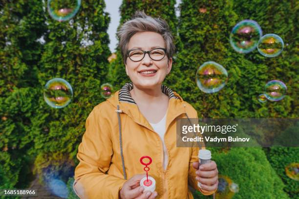 happy active mature woman blowing soap bubbles on green background - bubbles happy stockfoto's en -beelden