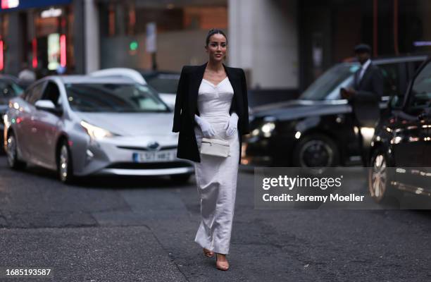Tamara Kalinic is seen wearing golden earrings, a black blazer with shoulder pads, underneath a white bustier maxi dress with structured chest area,...