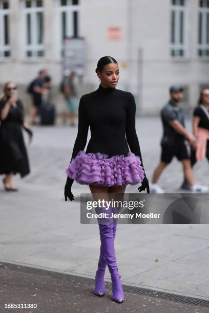 Ella Balinska is seen wearing silver hoop earrings, black sunglasses, a long-sleeved mini dress with black turtleneck and purple tulle tutu, black...