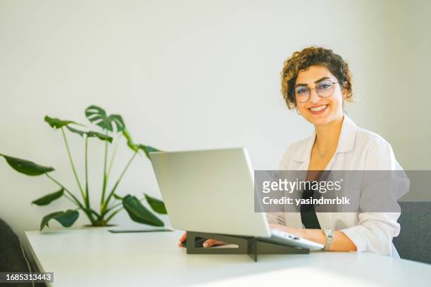curly haired woman in white uniform working from laptop in office - laptop netbook stock pictures, royalty-free photos & images