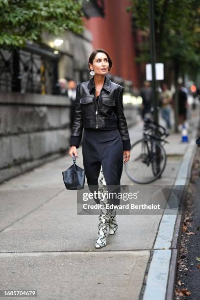 Geraldine Boublil wears earrings, a black leather jacket, a black midi slit skirt, a black leather bag, thigh high white pointed snake print high...