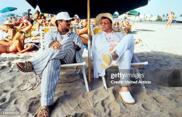 Robin Williams and Nathan Lane sitting under an umbrella on the sand at the beach in a scene from the film 'The Birdcage', 1996.