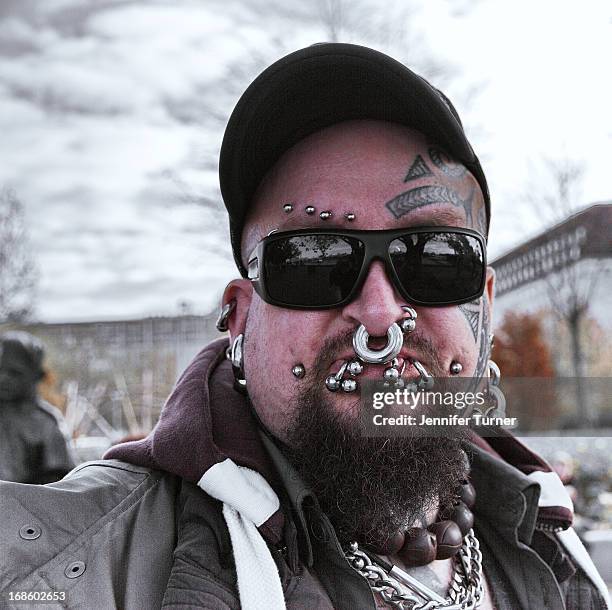 Candid portrait taken on the South Bank in London of a man wearing a baseball cap, sunglasses and who has lots of piercings in his eyebrows, nose,...