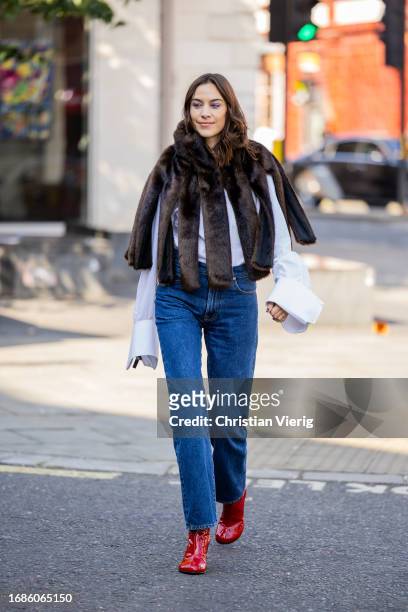 Alexa Chung wears fur cape with slits, denim jeans, white button shirt, red ankle boots outside JW Anderson during London Fashion Week September 2023...