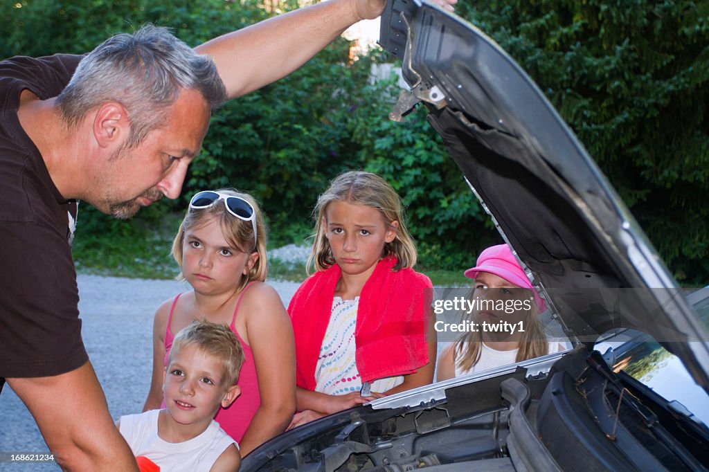 Dad trying to fix broken car while his kids wait sadly