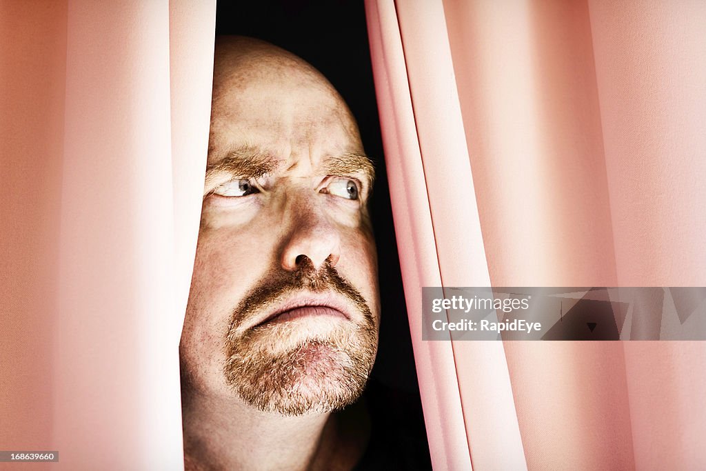 Angry man frowns, peering through closed curtains: noisy neighbours?